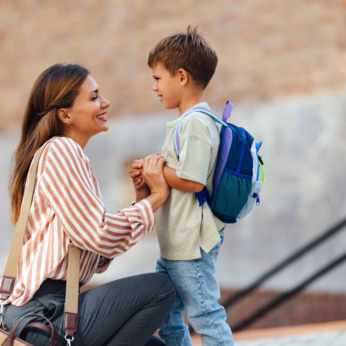 A mother lovingly sends her young son off on his first day at elementary school. The child wears a backpack, and they share a joyful moment outdoors.