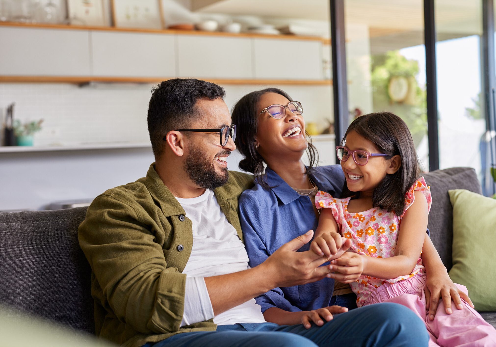 Cheerful middle eastern family with eyeglasses of three having fun together at home. Mixed race parents talking with their little girl in living room. Cheerful family with specs embracing and playing with beautiful indian daughter girl in arms.
