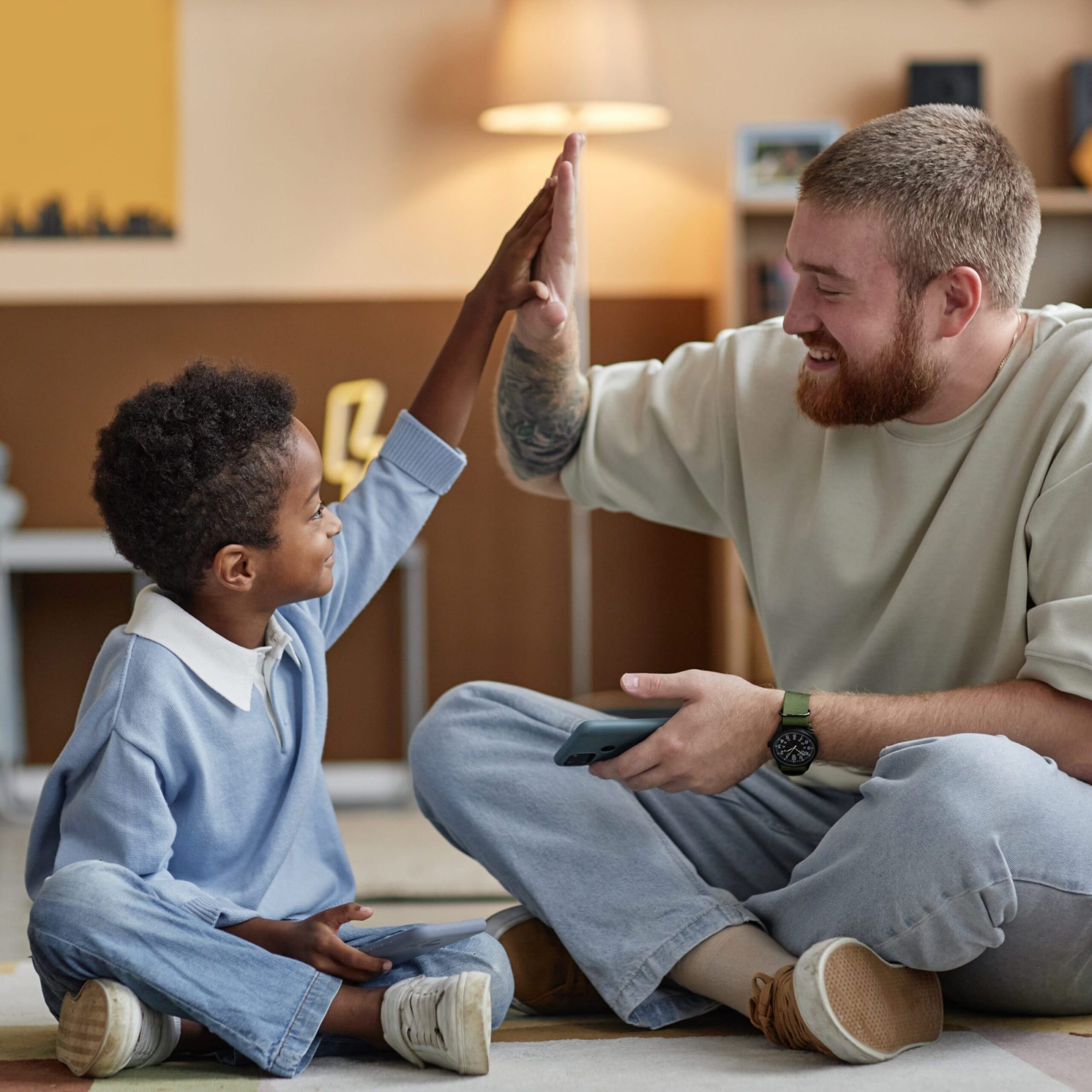Little smiling Black boy giving high five to joyful adoptive father while celebrating winning at mobile phone game sitting on floor in kids room
